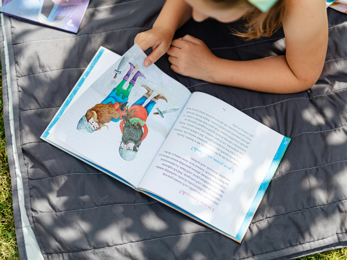 Child reading a colourful storybook outdoors on a picnic blanket.