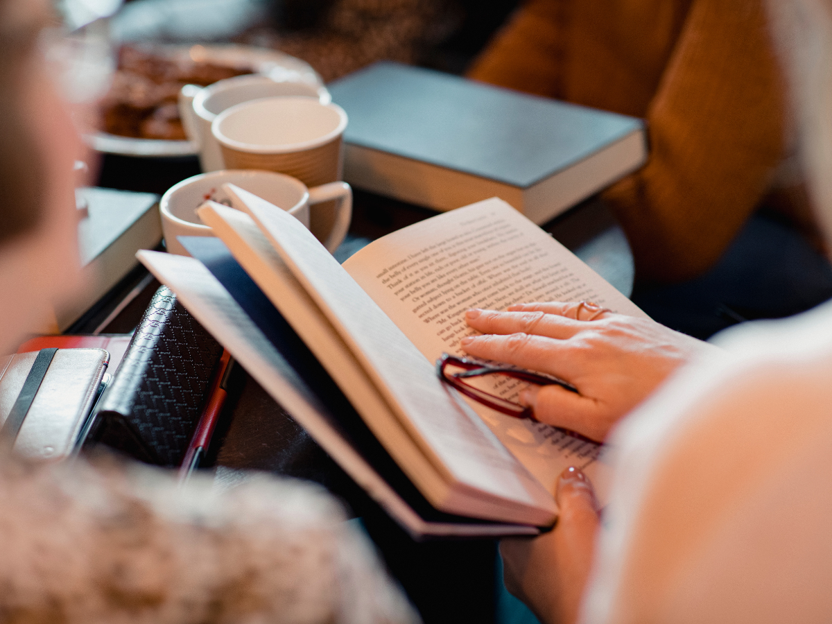 Person reading an open book at a table with drinks nearby.
