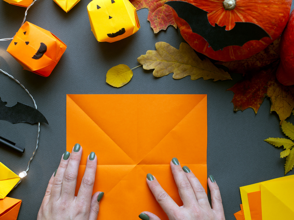 Hands folding orange paper surrounded by autumn-inspired craft decorations.